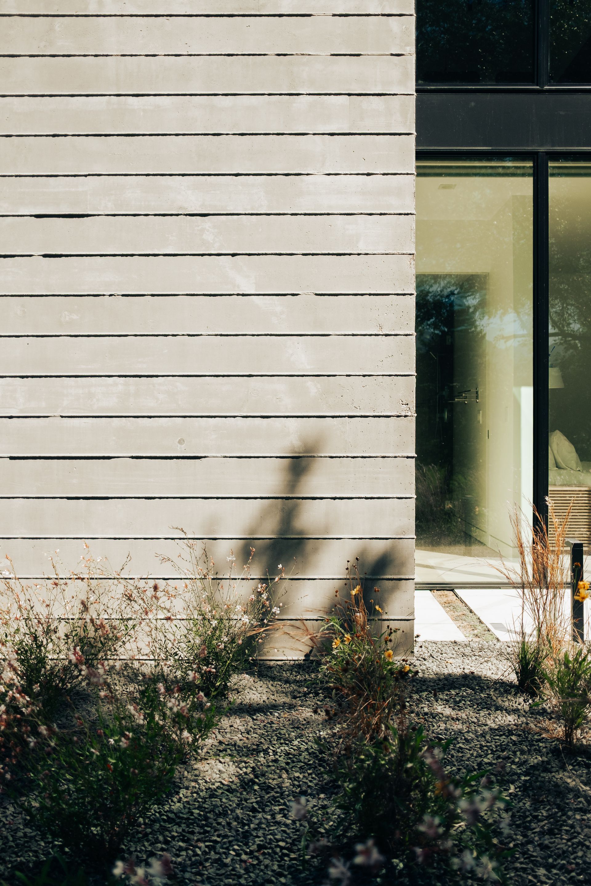 Concrete wall with horizontal grooves next to a glass window and small plants in a gravel garden.