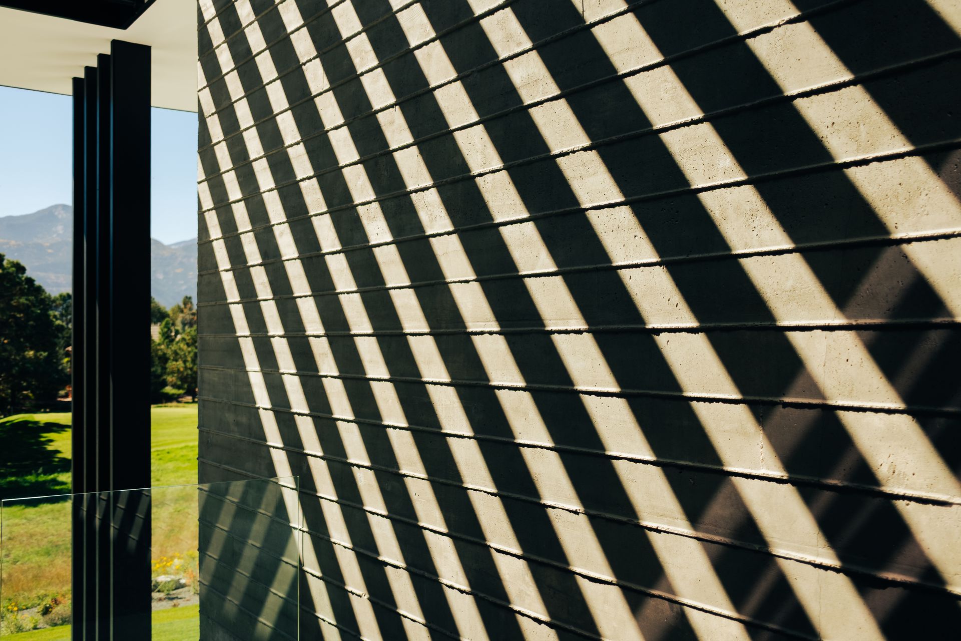 Shadows from vertical slats create diagonal patterns across a textured concrete wall, overlooking a sunny landscape.