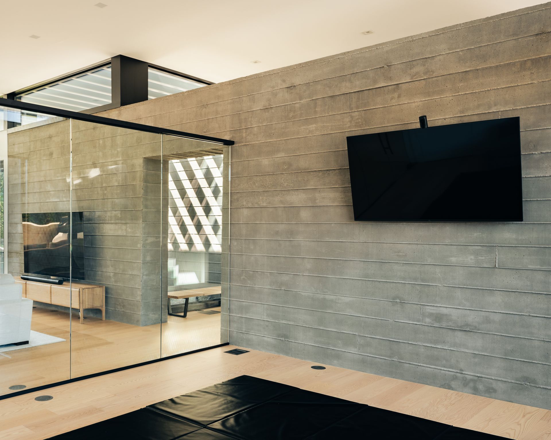 A black flat-screen television is mounted on a textured gray concrete wall in a room with light wood floors and glass walls.