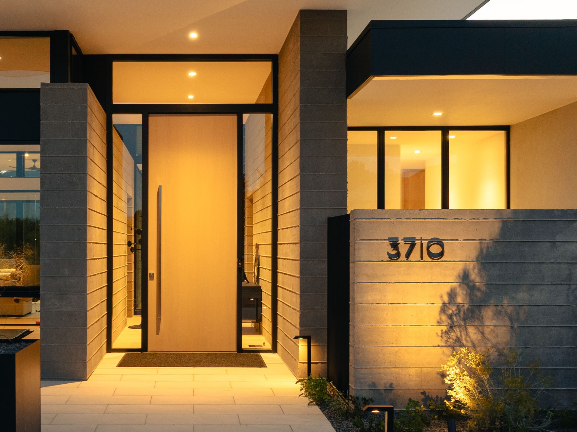 A modern home entrance with a light wood door, glass side panels, stone accents, and the address 3710 lit at night.