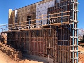 A construction site shows a reinforced concrete wall being framed with metal panels and scaffolding under a clear blue sky.