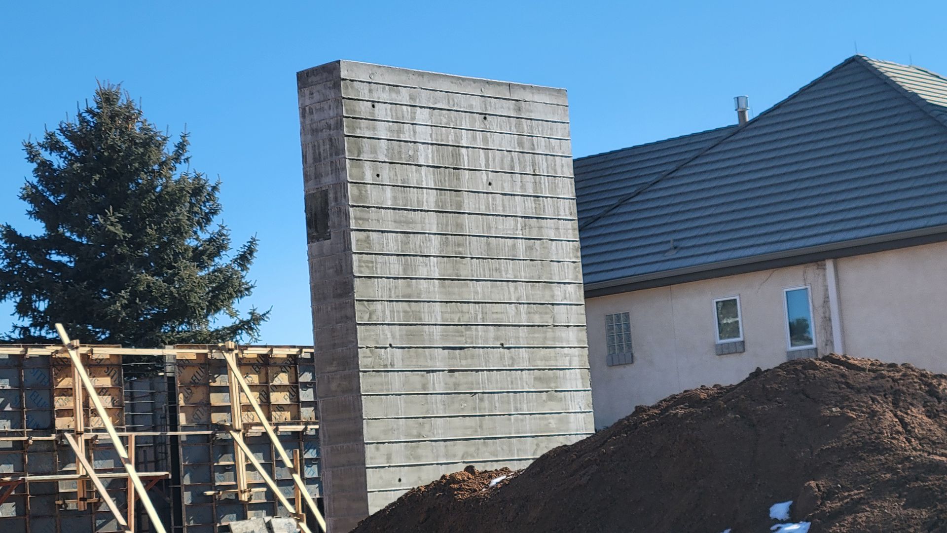A tall, rectangular concrete structure stands next to a house under construction and a pile of dirt against a blue sky.