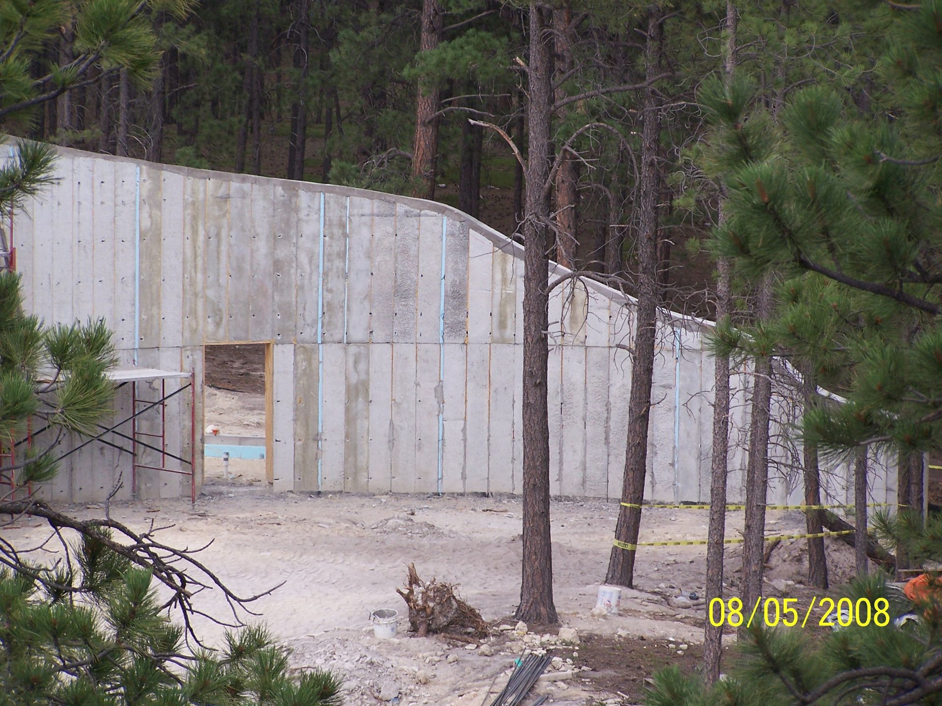 A tall, sloped concrete foundation wall stands in a forest setting, featuring a doorway opening and scaffolding nearby.