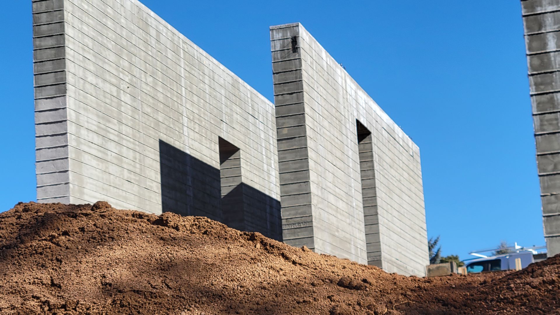 Concrete foundation walls for a building under construction, standing above a mound of brown earth against a clear sky.