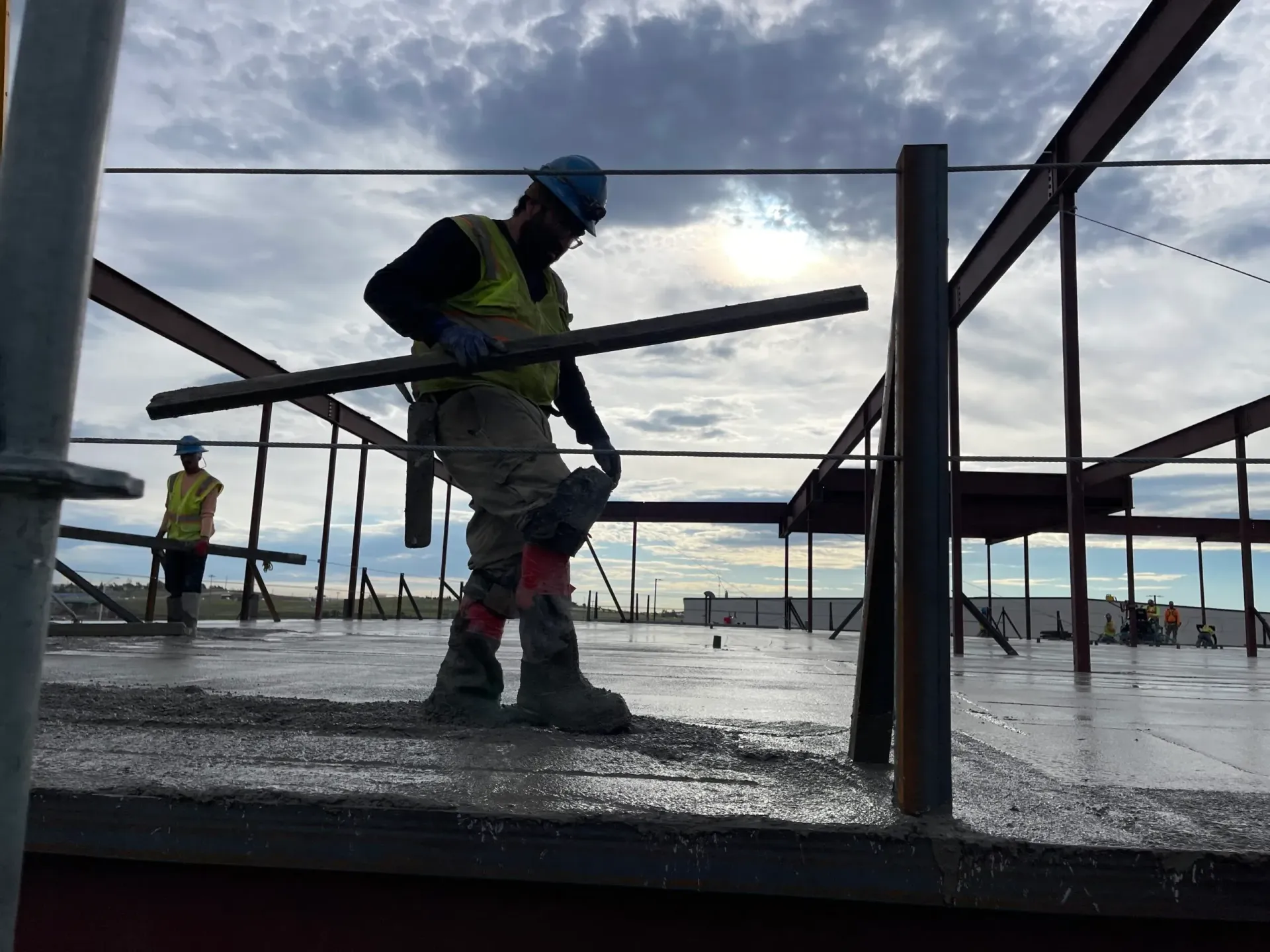 Construction workers in hard hats and safety vests working on a wet concrete floor within a steel frame structure.