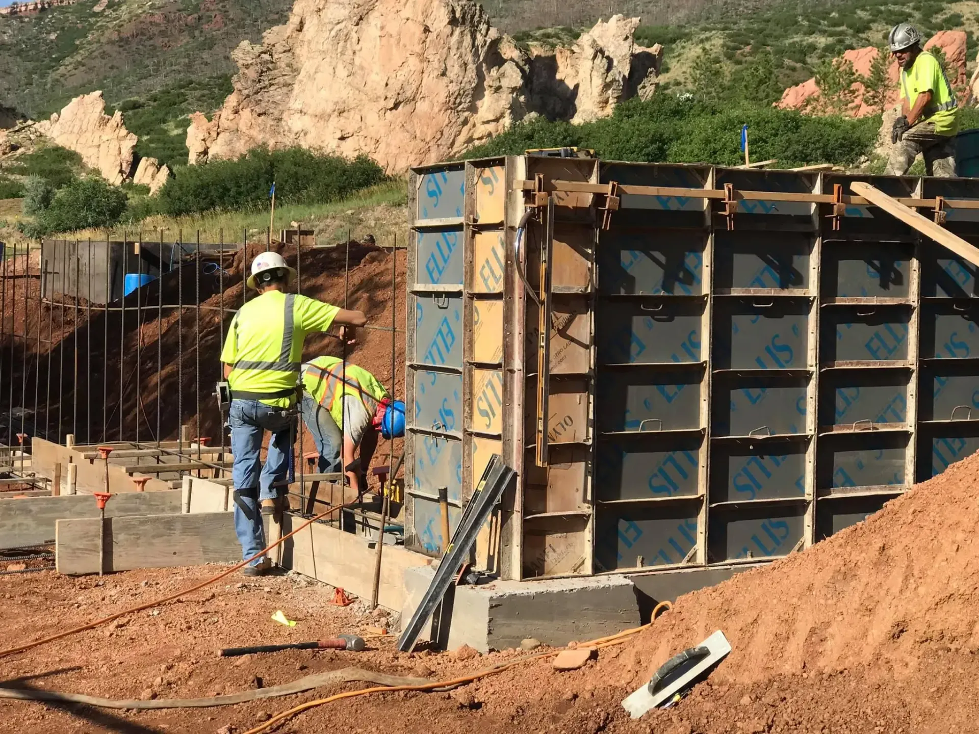 Construction workers in high-visibility gear assemble concrete formwork at a site with rocky hills in the background.