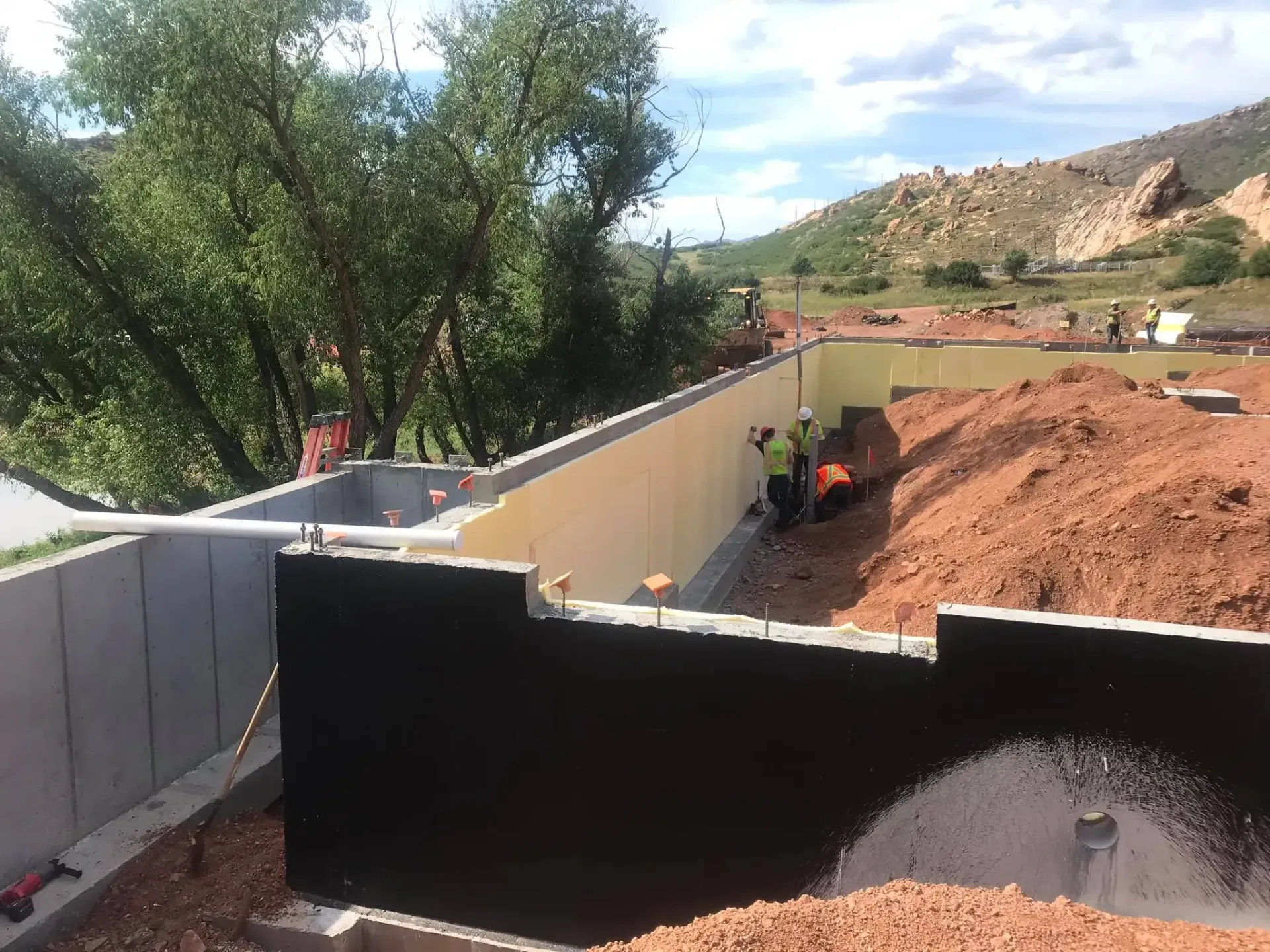 Construction workers in a partially built concrete foundation with black waterproofing on the exterior basement walls.