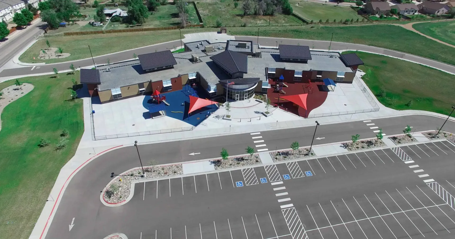 Aerial view of a school building with a large parking lot, paved playground areas, and colorful shade structures.