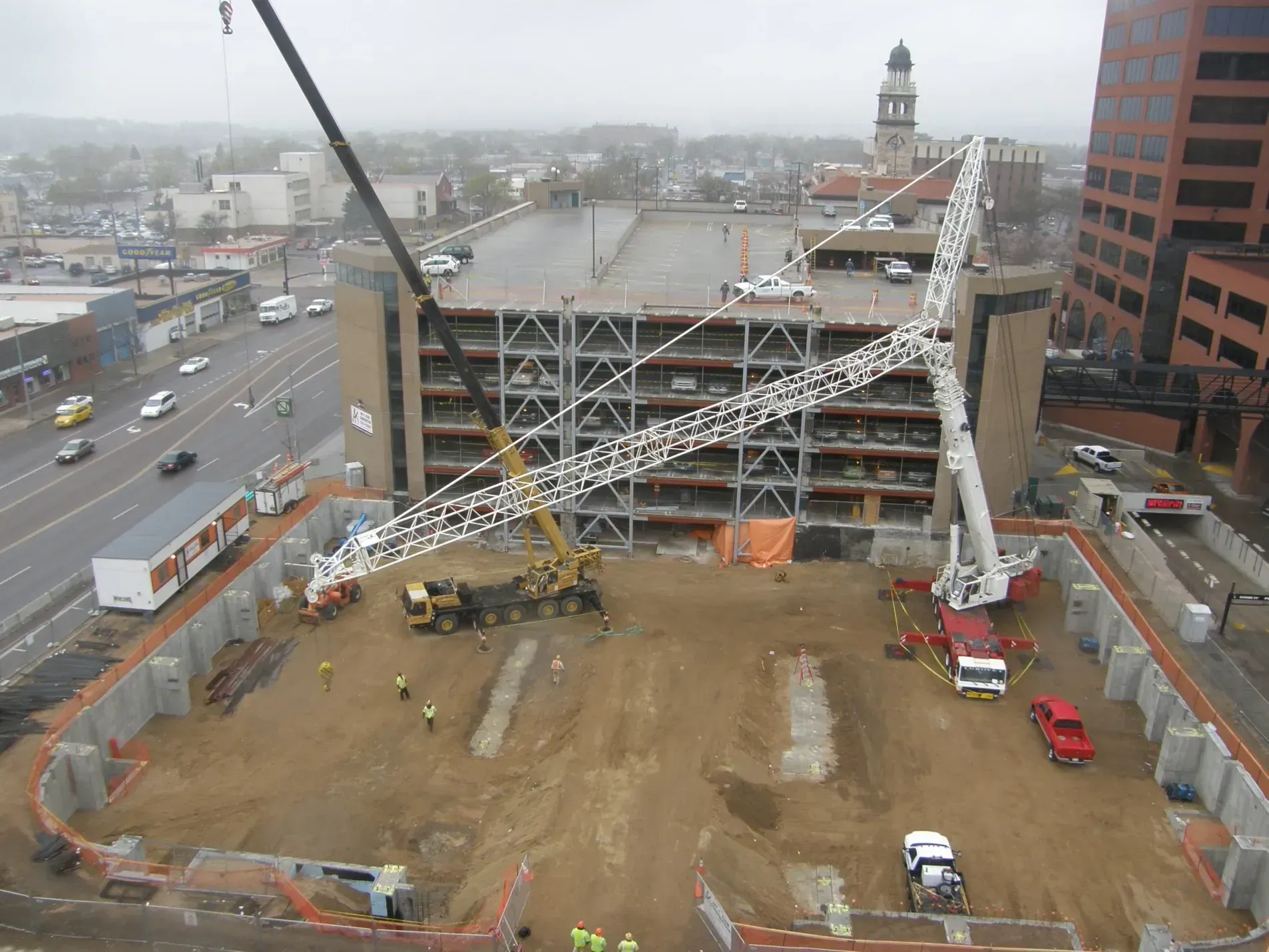 Two cranes working in a dirt construction site next to a multi-story parking garage and city buildings.