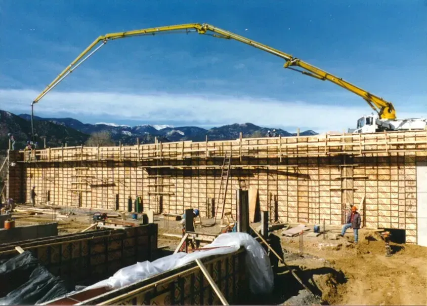 A yellow concrete pump truck extends its boom arm over a large wooden construction wall against a blue sky.