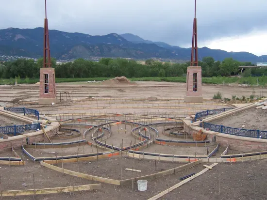 Construction site with curved concrete forms, two brick columns topped with spires, and mountains in the background.