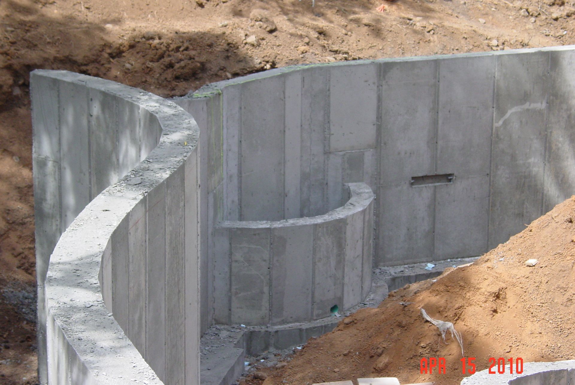 Curved concrete walls of a basement foundation under construction with dirt mounds nearby.