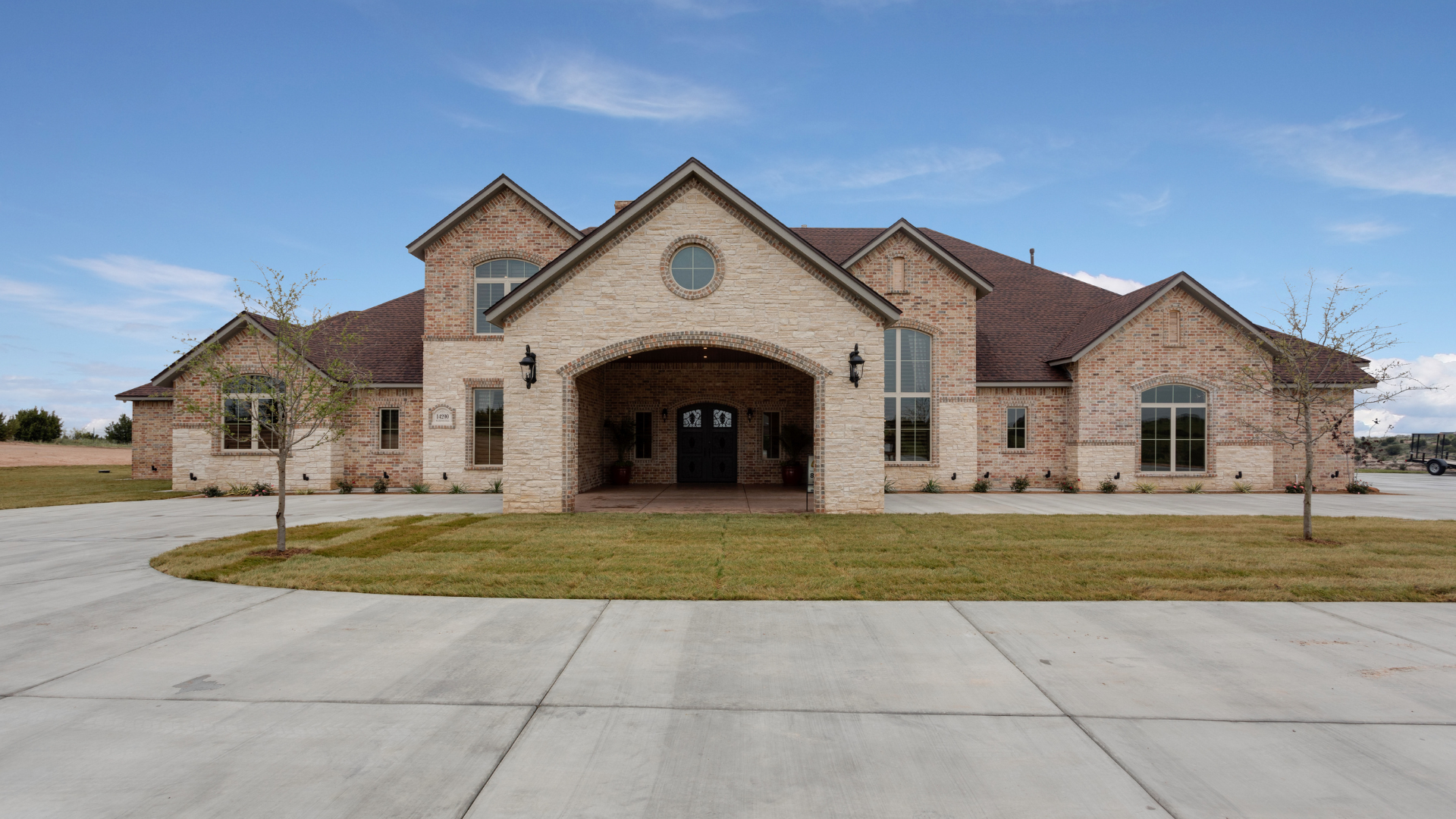 A single-story, light-colored brick house with a dark gabled roof, an arched entryway, and a large concrete driveway.
