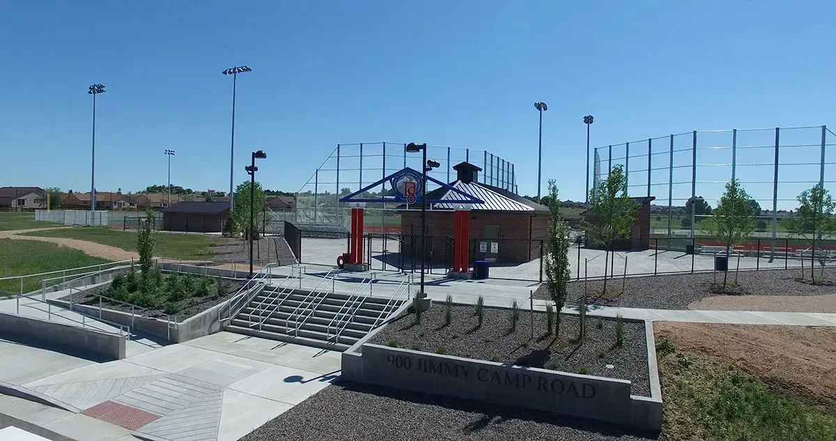 Outdoor park area featuring a small, open-sided pavilion, landscaped garden beds, and sports netting under a blue sky.