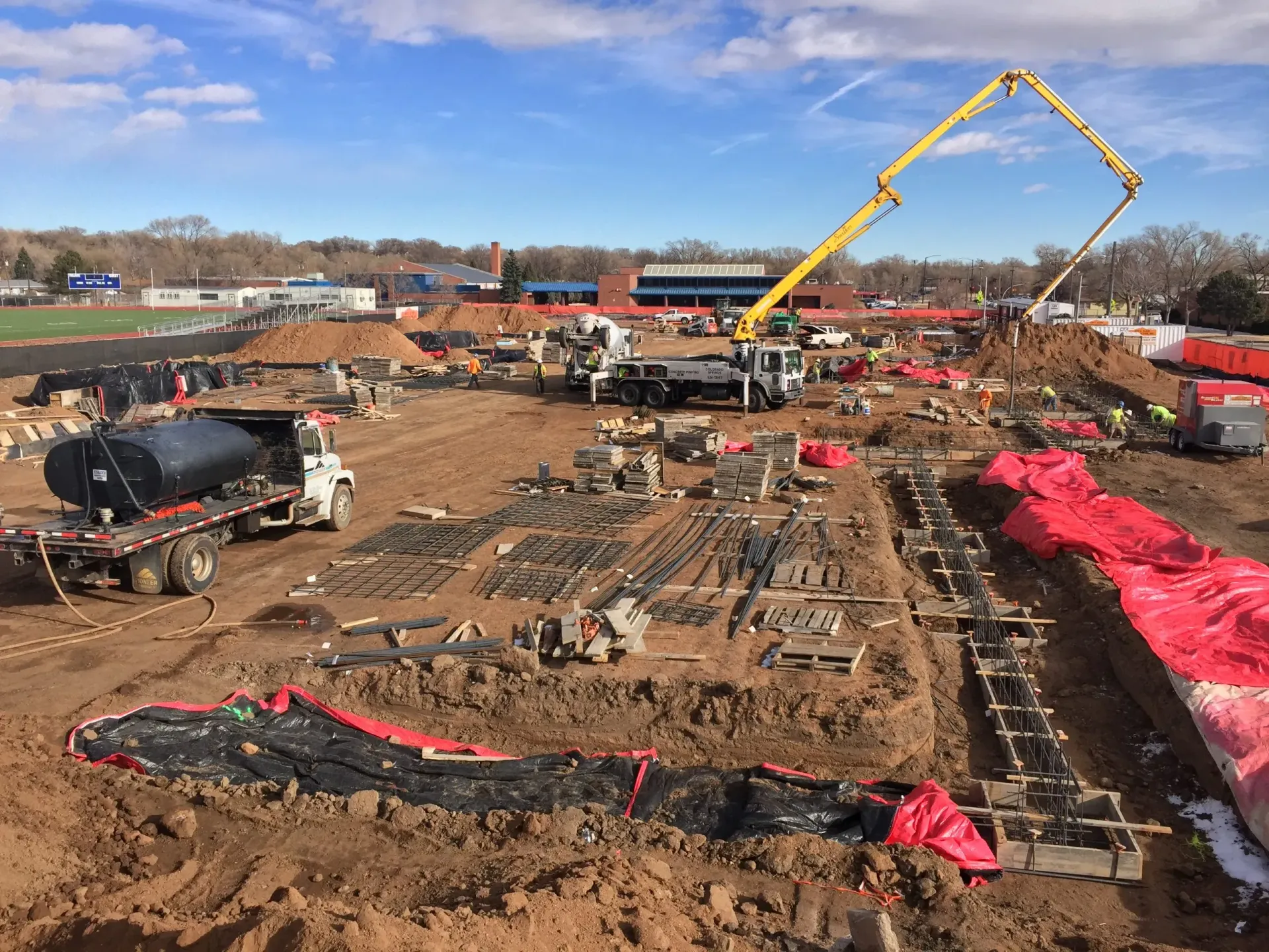 Construction site with a concrete pump truck, a fuel tanker, and trenches with rebar, under a sunny sky.