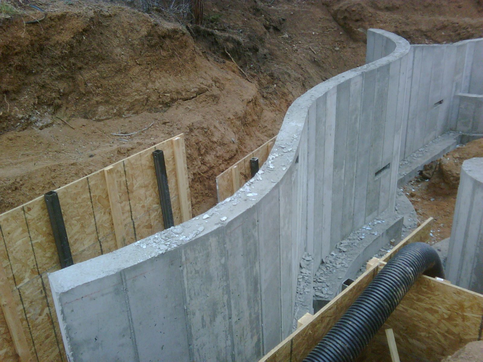 Curved concrete foundation walls under construction in an excavated trench with wooden forms and drainage pipe.