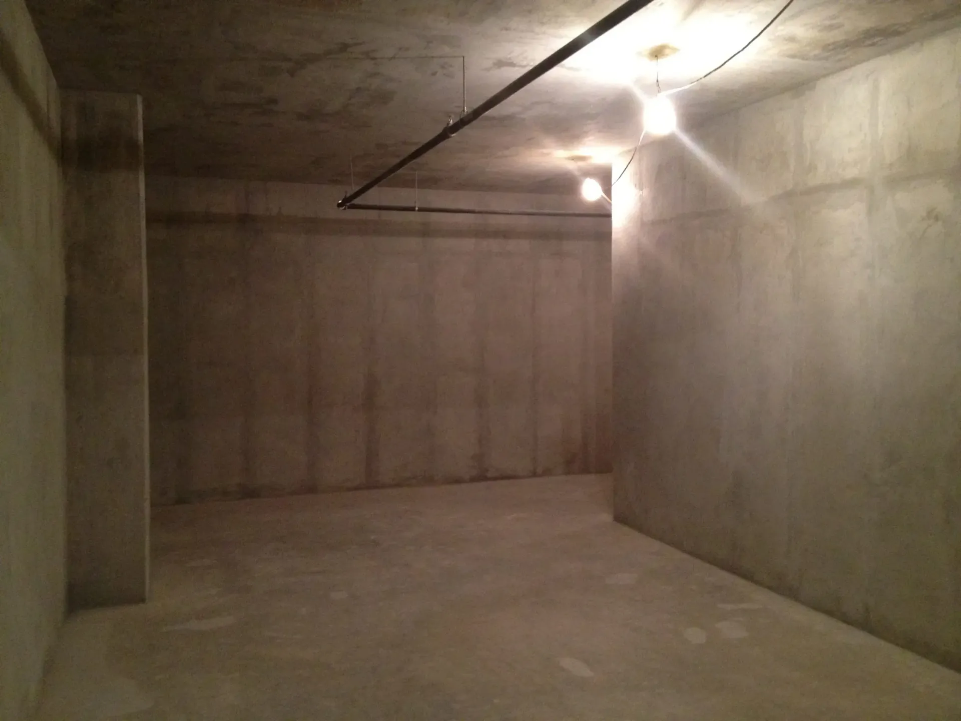 An empty, dimly lit concrete basement room with exposed light bulbs and a pipe running across the ceiling.