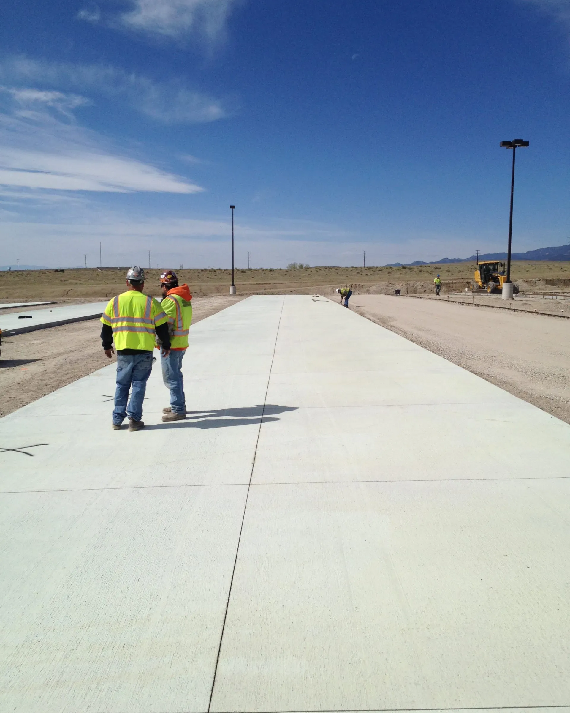 Two workers in high-visibility vests stand on a freshly poured concrete slab in an open desert environment.