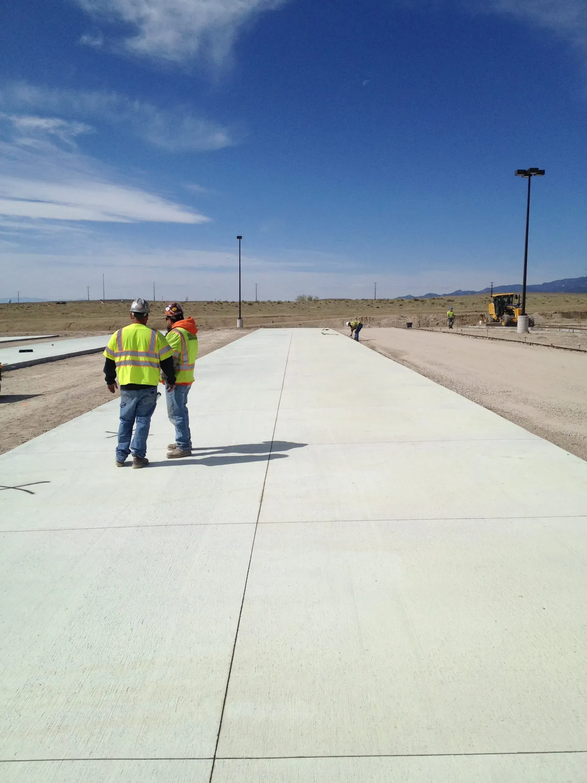 Two workers in high-visibility safety vests stand on a freshly poured concrete road in an open, desert landscape.