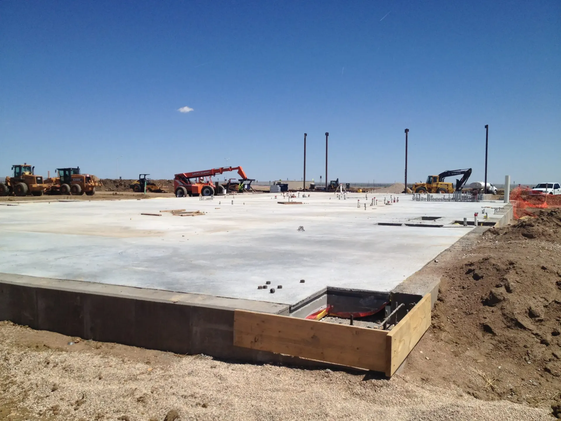 A wide shot of a concrete building foundation under construction in an open, dirt-filled field under a clear blue sky.