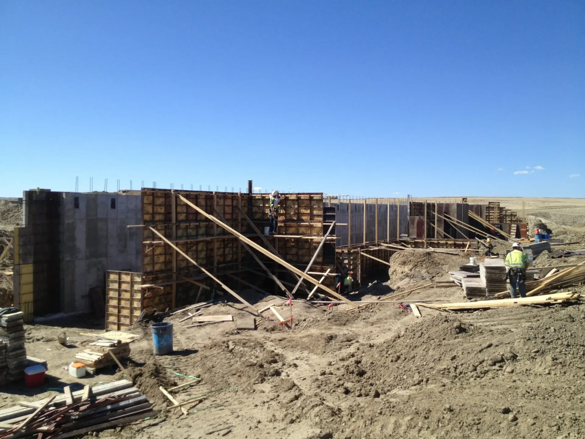 Construction workers erect modular concrete forms for a wall in a dry, open field under a clear blue sky.