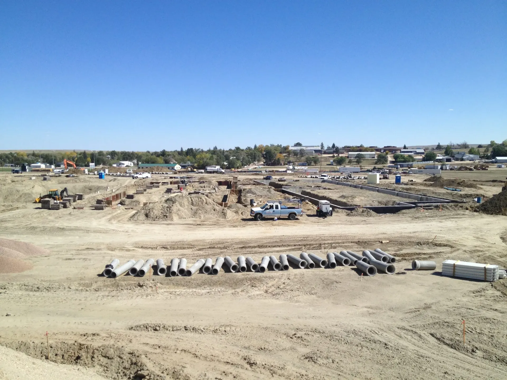 Construction site with excavated earth, concrete pipes lined up in the foreground, and equipment under a clear blue sky.