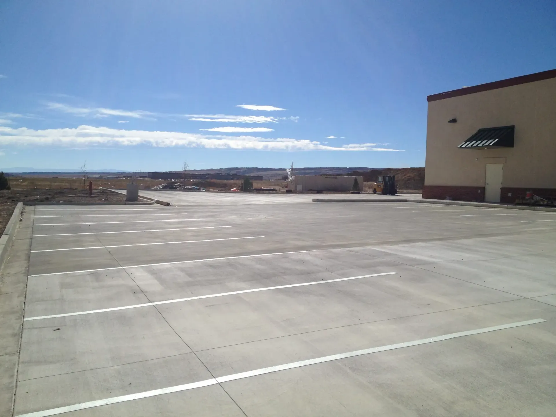 A newly paved concrete parking lot with painted white parking lines next to a tan commercial building under a blue sky.