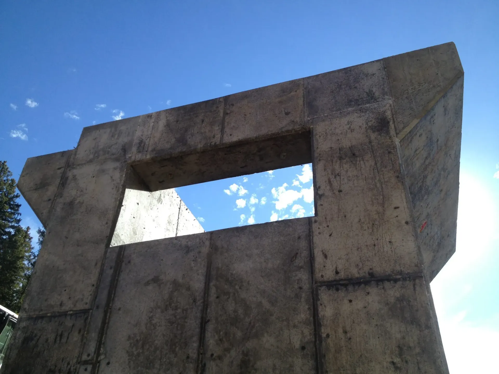 Low-angle view of a weathered concrete structure with a rectangular cutout against a bright blue, cloudy sky.