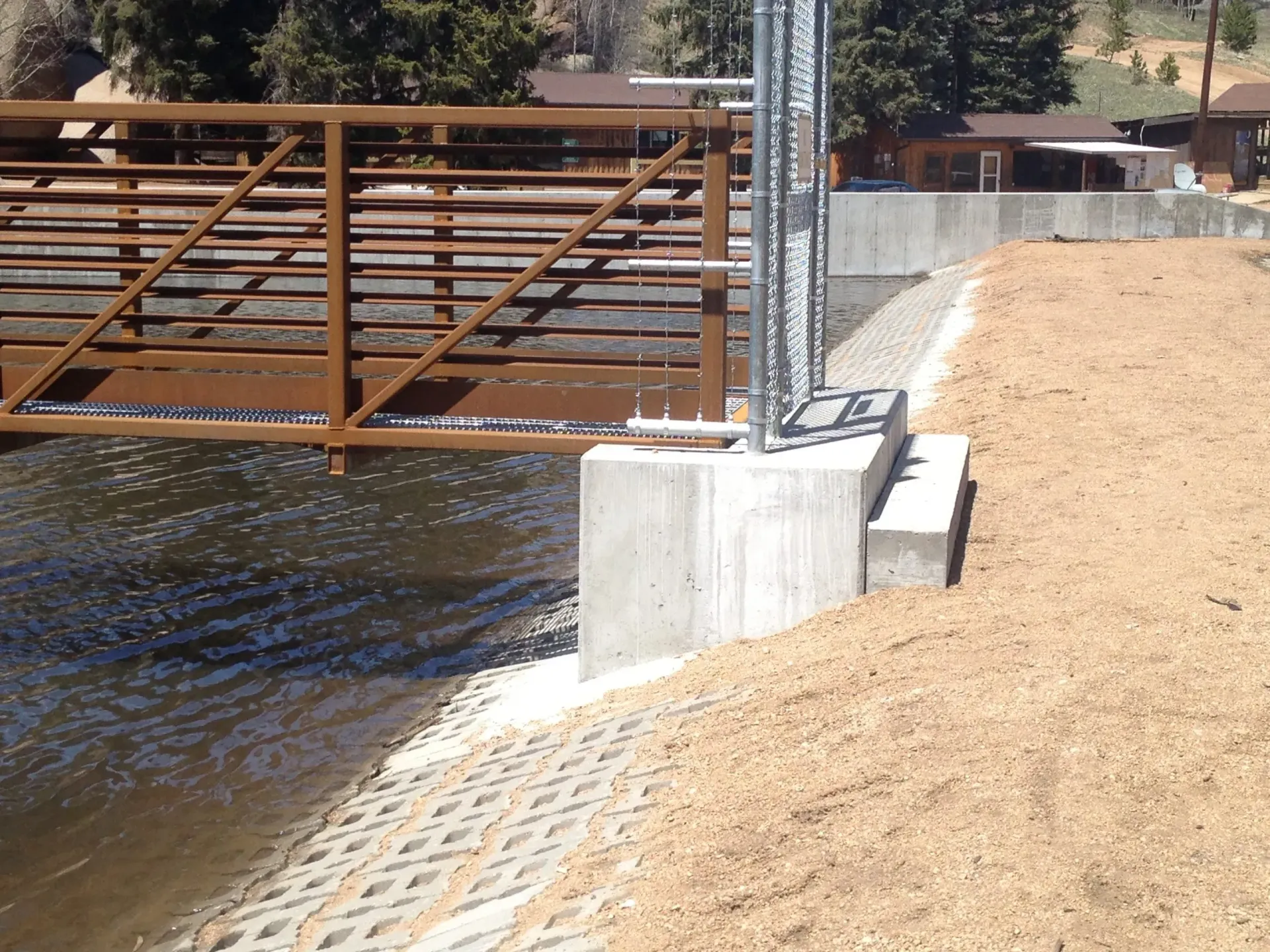 A metal pedestrian bridge spans a river, attached to a concrete abutment next to a sloped embankment with soil and stones.