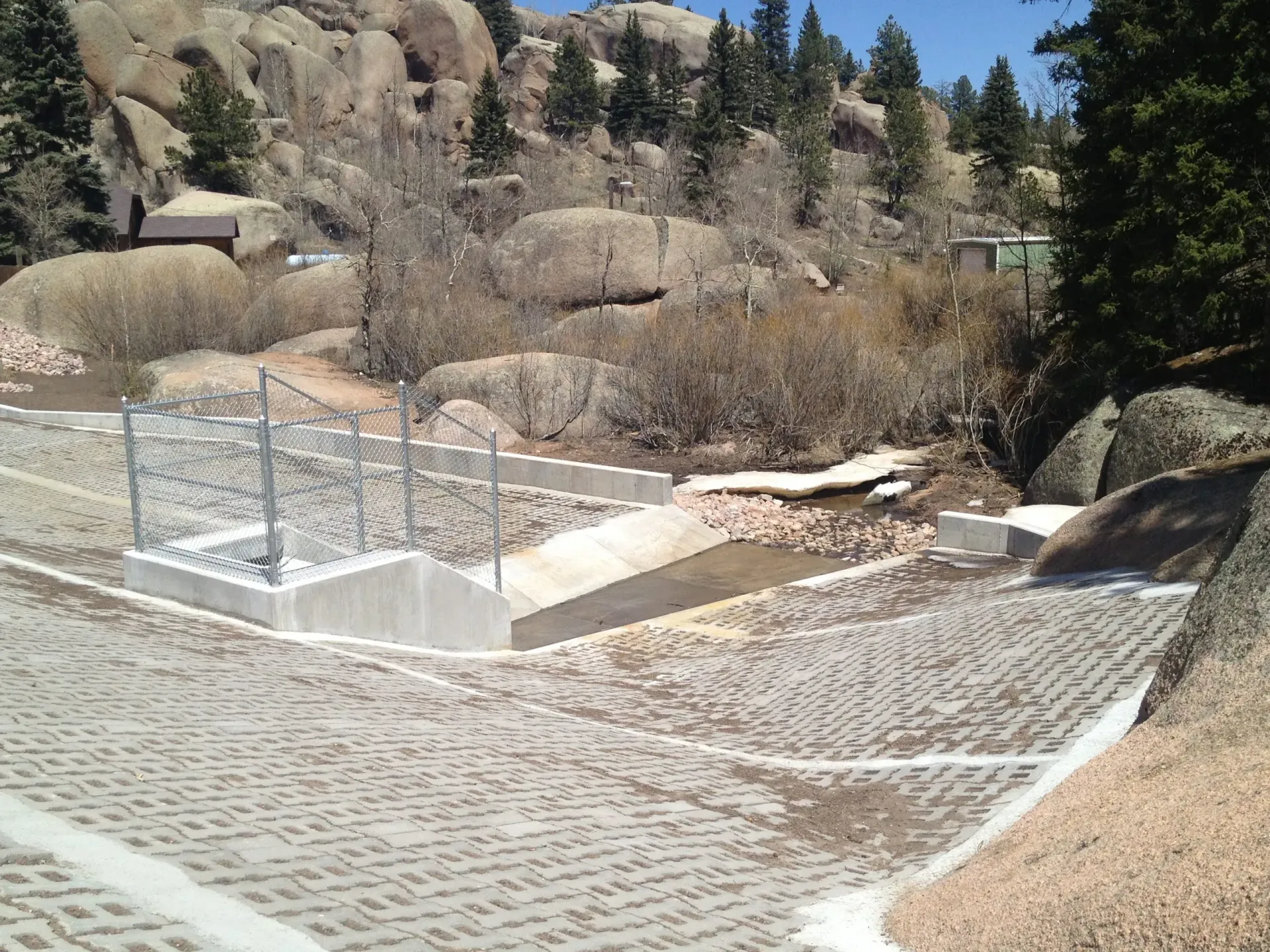 A concrete drainage channel with a metal grate and interlocking pavement blocks in a rocky, forested area.