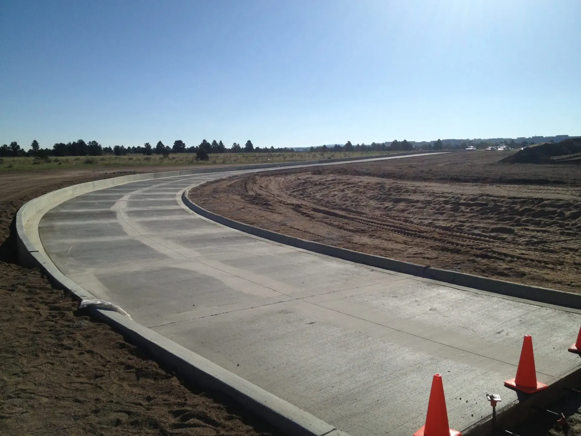 Newly paved curved concrete road bordered by curbs in a construction site under a clear blue sky.