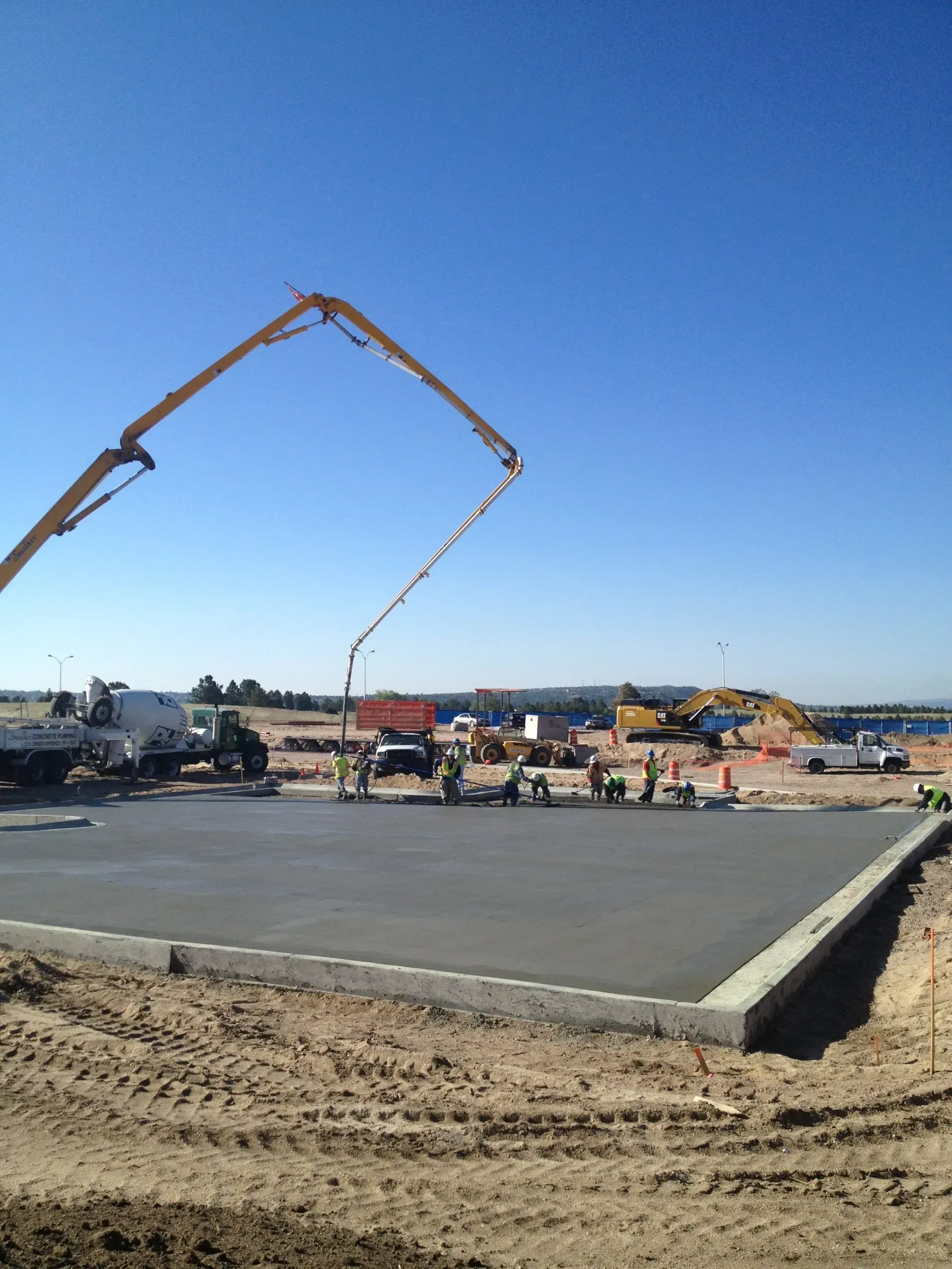 Construction workers finish a large concrete slab with a concrete pump truck extending over it under a clear blue sky.