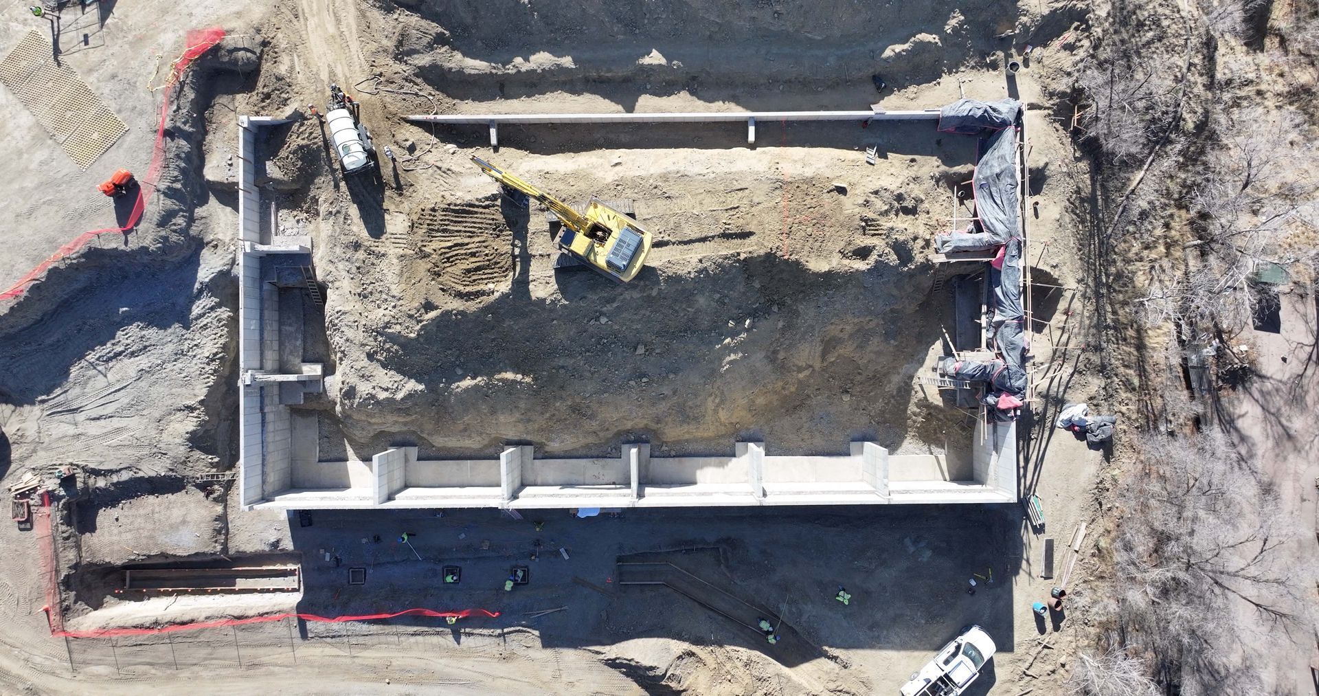 Aerial view of a rectangular concrete building foundation under construction with an excavator inside the site.