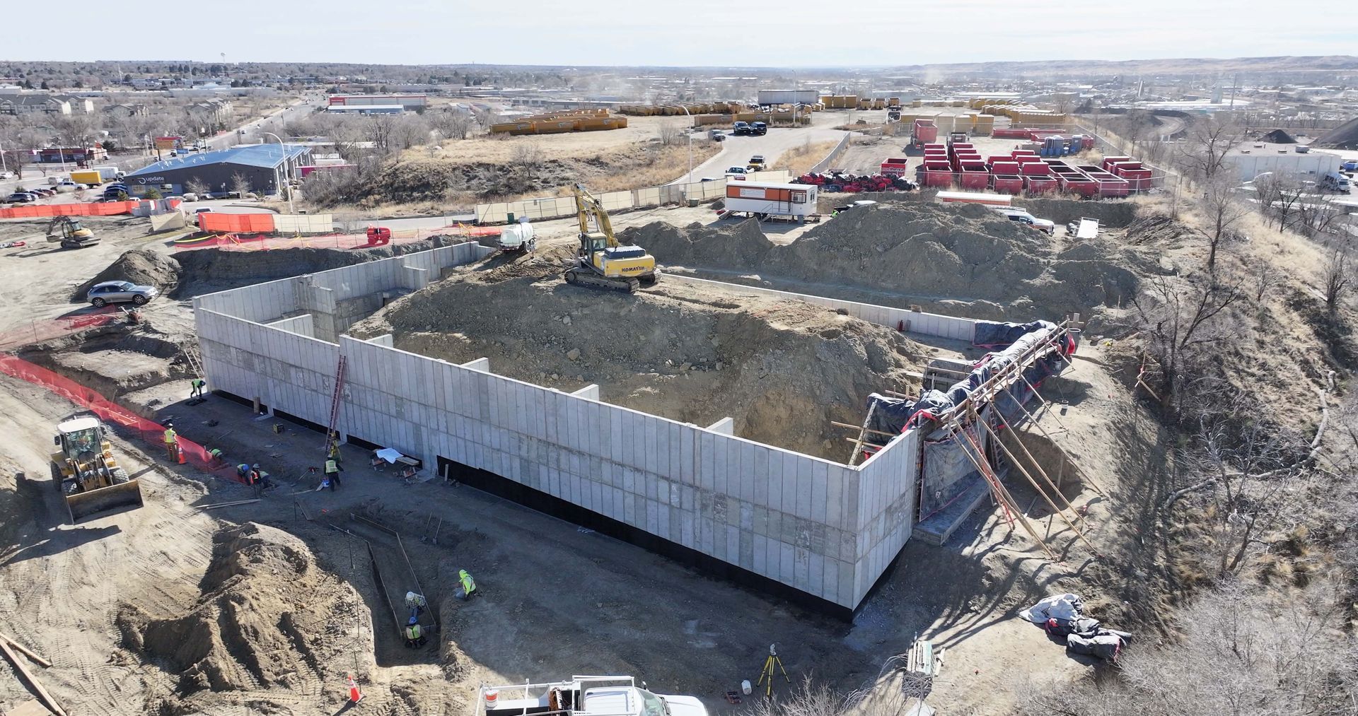 An aerial view of a construction site featuring a large, rectangular concrete foundation wall filled with dark soil.