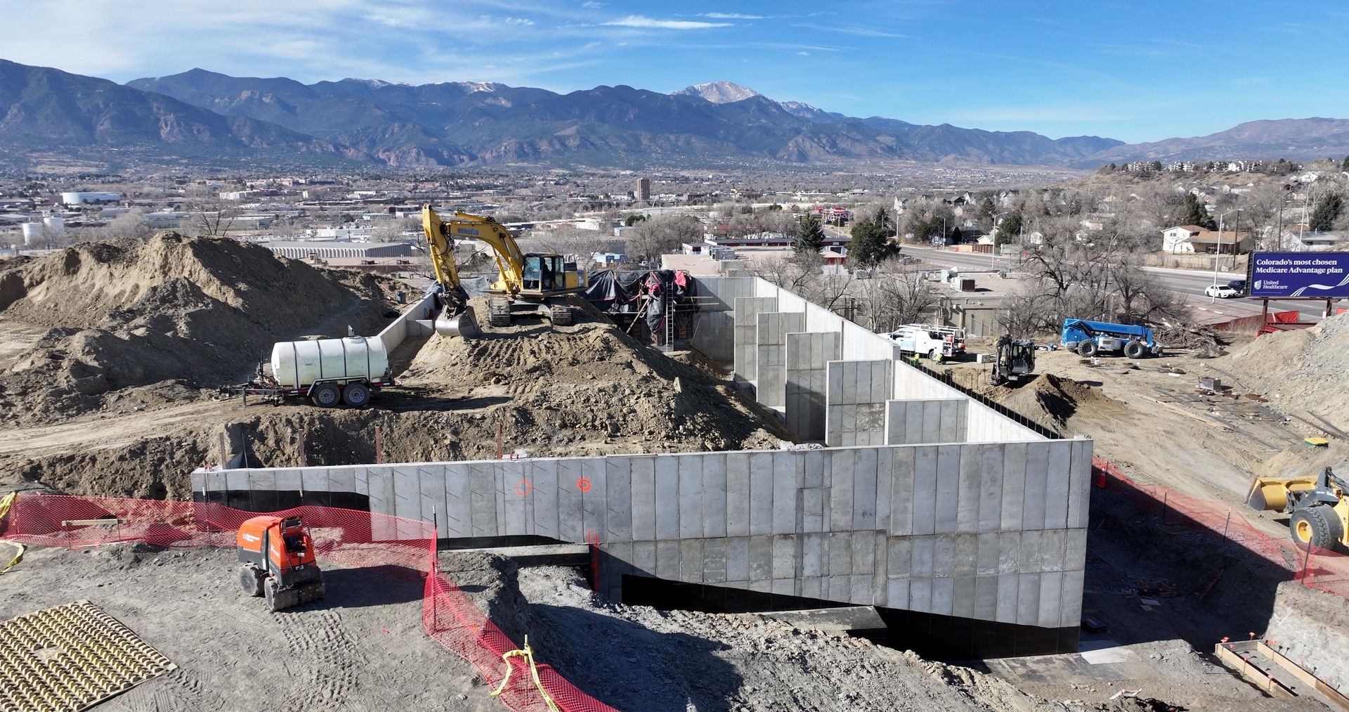 Aerial view of an excavator working near a large, L-shaped concrete retaining wall under construction with mountains.