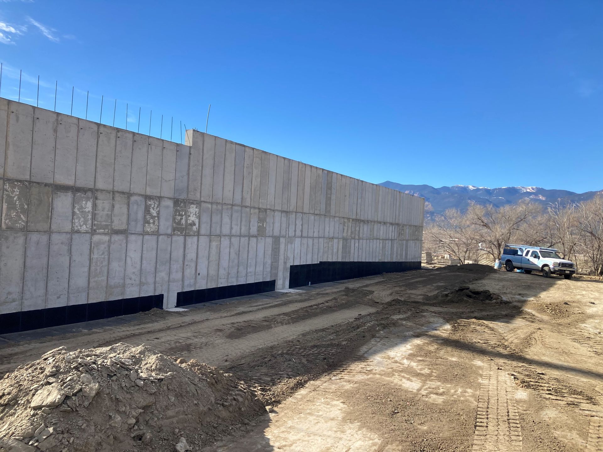 A long concrete foundation wall stands under a clear blue sky, with a white truck parked nearby on a dirt construction site.
