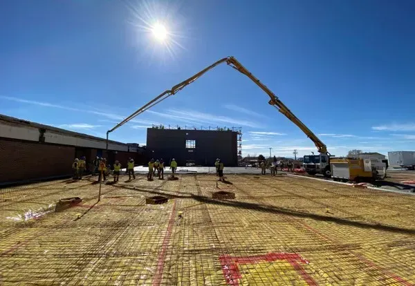 Construction workers on a site with a concrete pump boom extending over a grid of rebar and yellow vapor barrier.