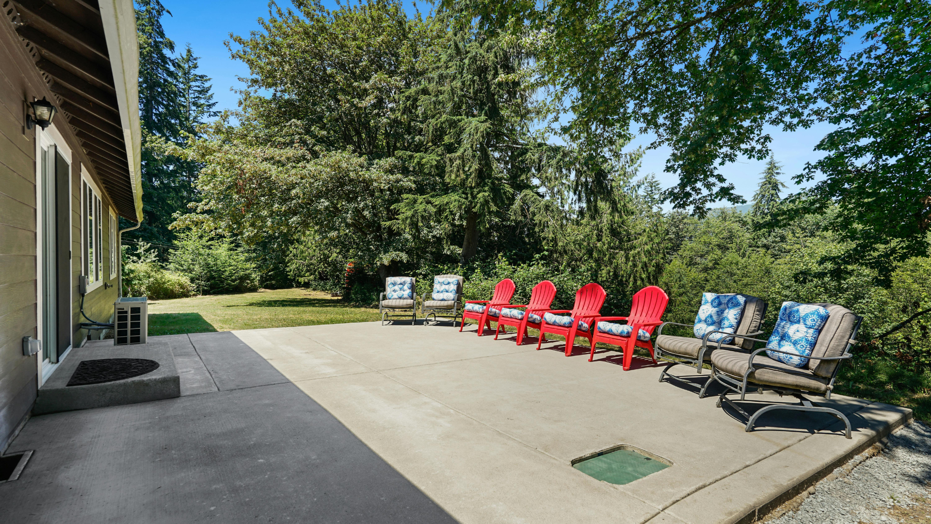 A concrete patio next to a house with red chairs and two outdoor cushioned seats facing a wooded backyard.