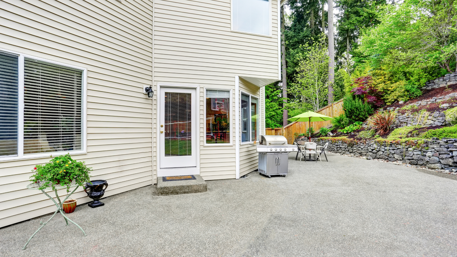 A backyard patio with a stone wall, a grill, an umbrella-shaded seating area, and a beige house with a glass door.