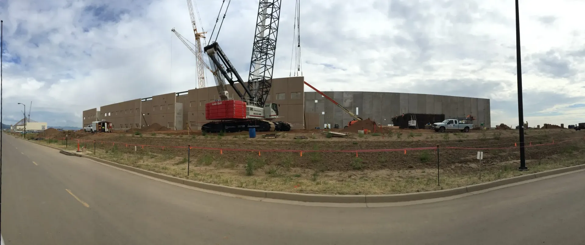 A large red crane lifts panels for a partially constructed concrete building under a cloudy sky.