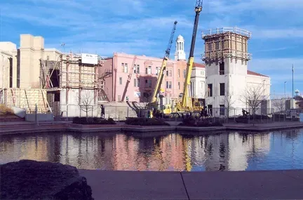 Construction equipment and cranes working near a partially renovated building by a water feature under a blue sky.