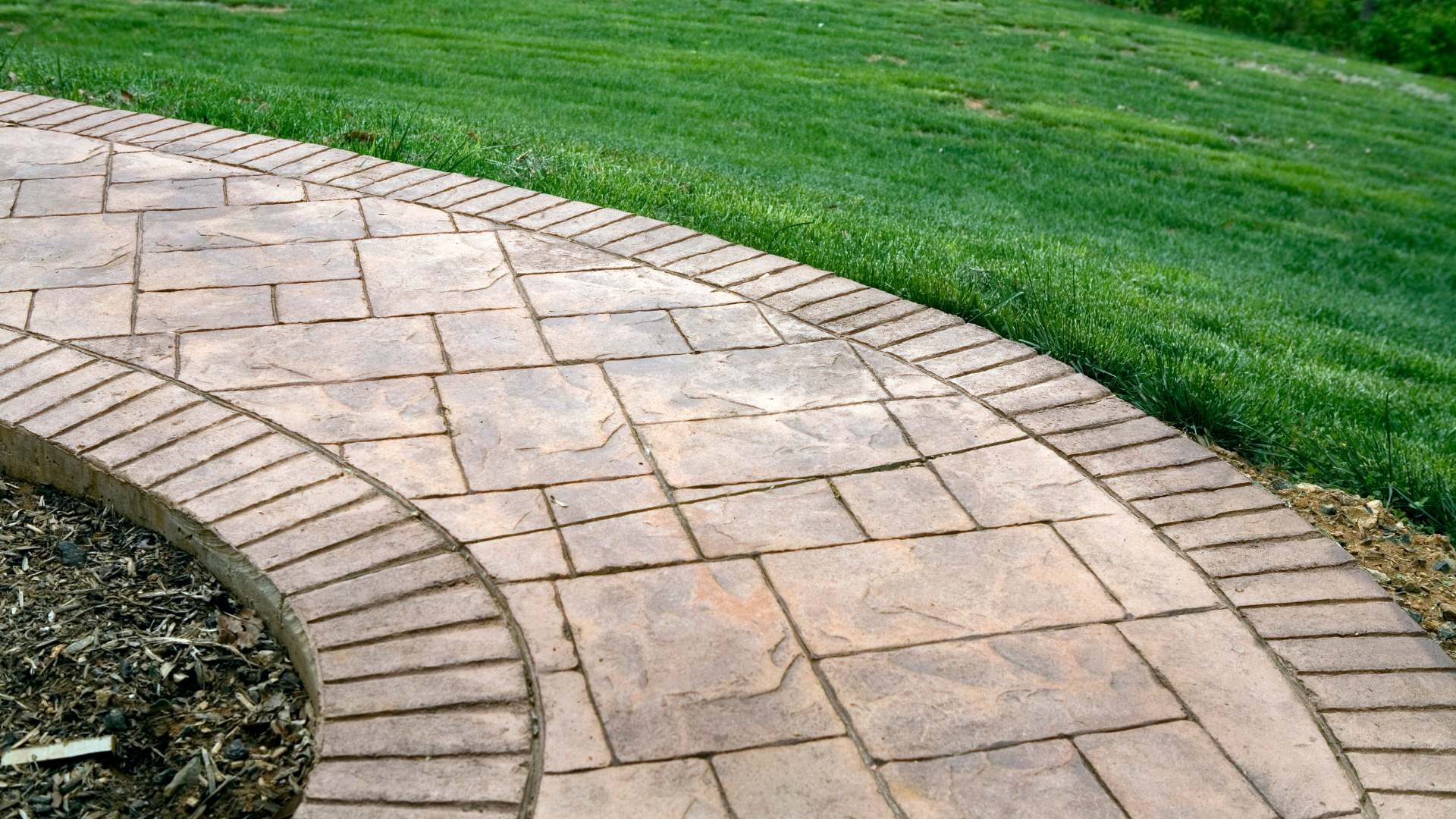 A curved stone paver walkway with a brick border, bordered by a dark mulch garden bed and vibrant green grass.