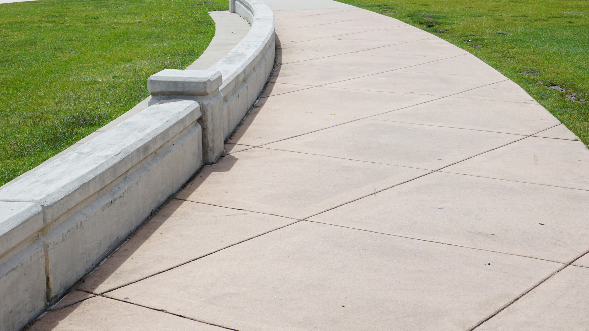 A curved concrete sidewalk lined with a low stone wall, bordered by green grass on both sides.