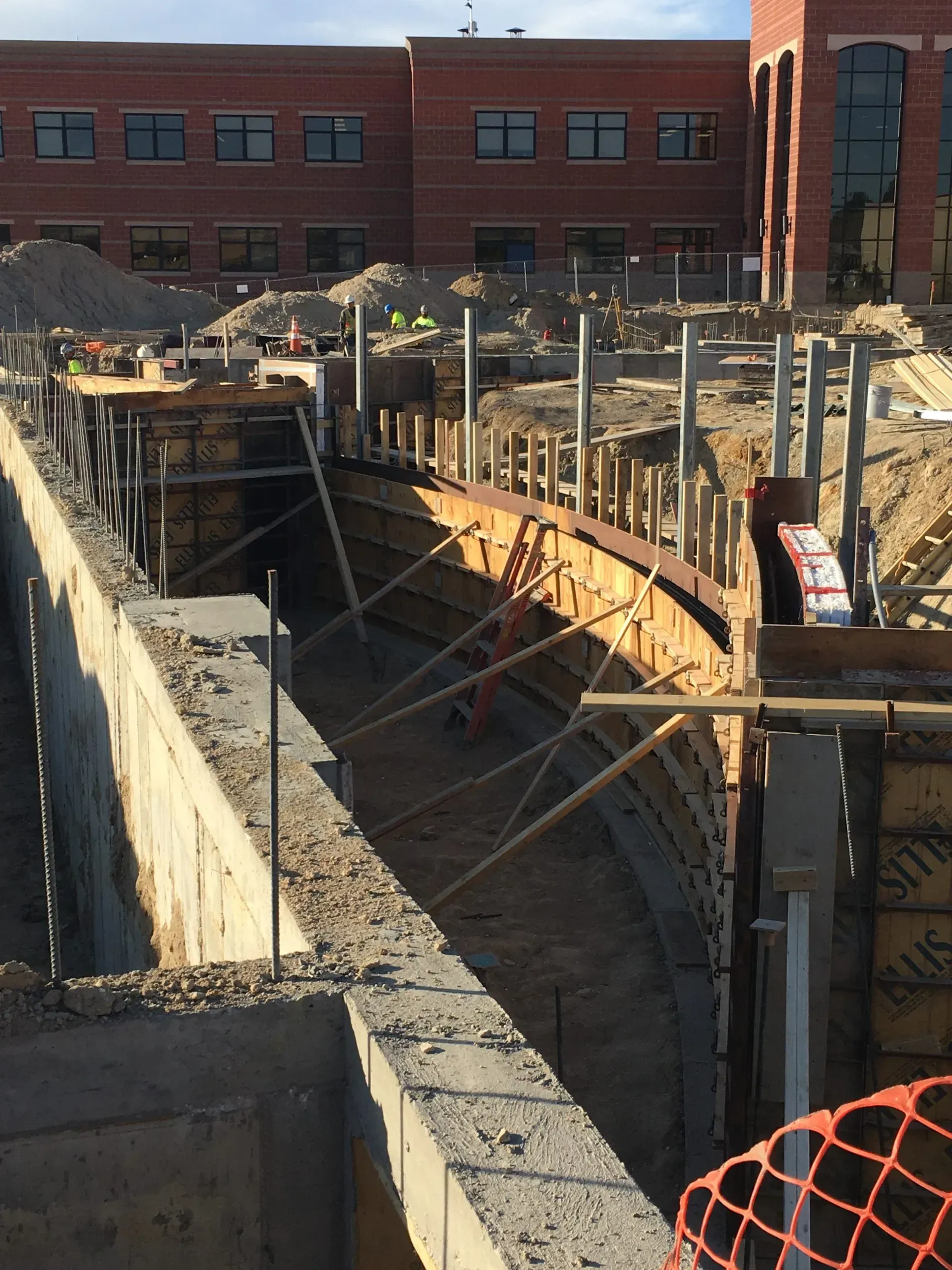 A construction site showing a curved concrete foundation formwork being installed in front of a brick building.
