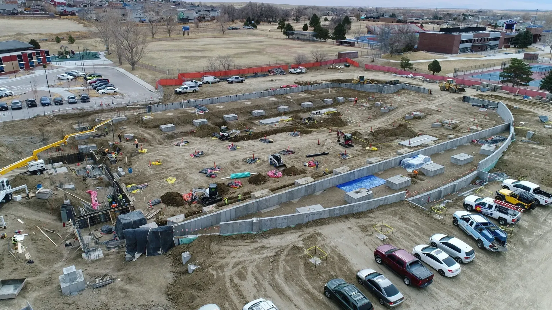 Aerial view of a construction site with a large, oval-shaped concrete foundation wall surrounded by parked vehicles.