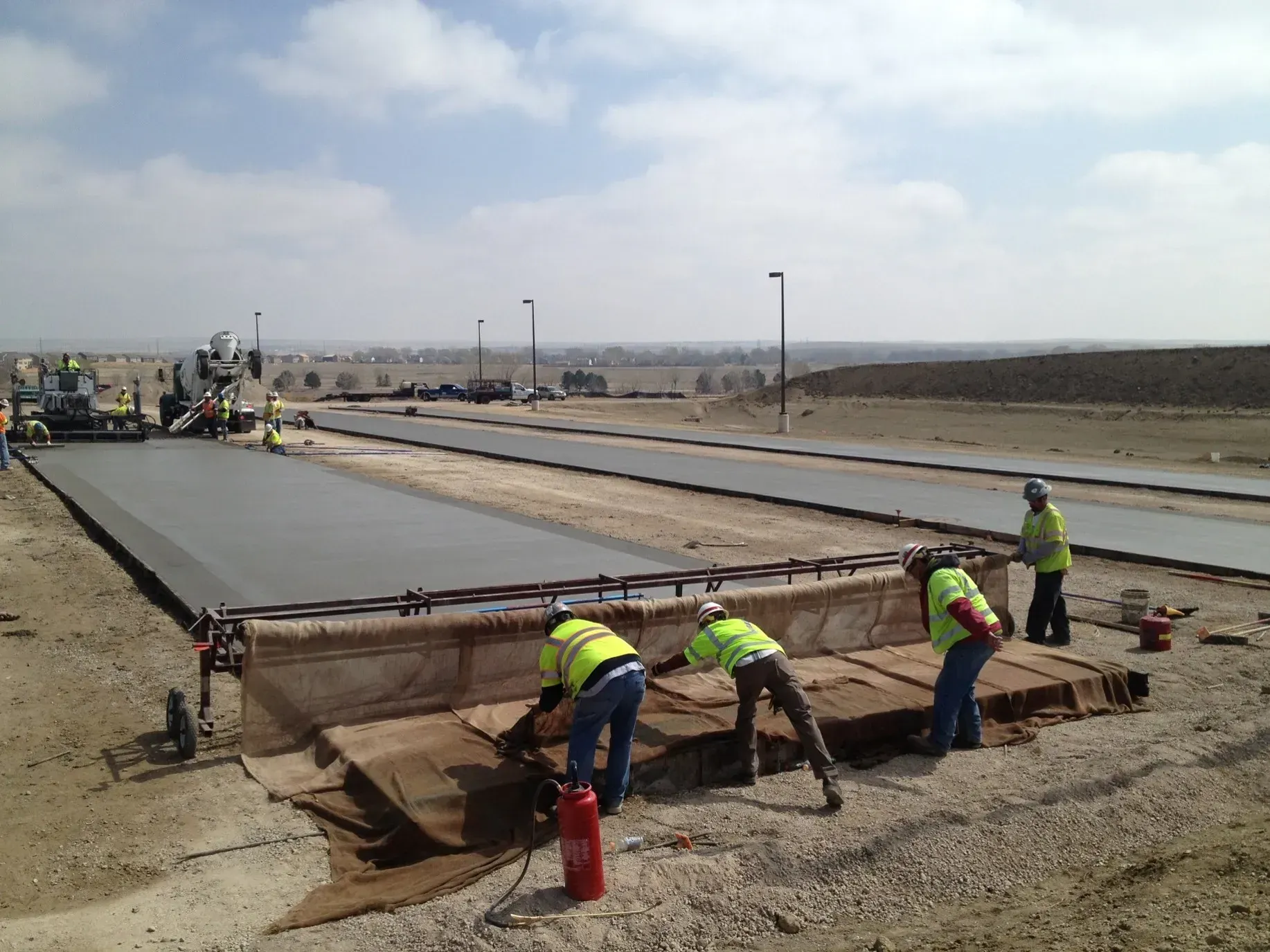Construction workers in high-visibility vests finish a newly poured concrete road section under a bright, cloudy sky.
