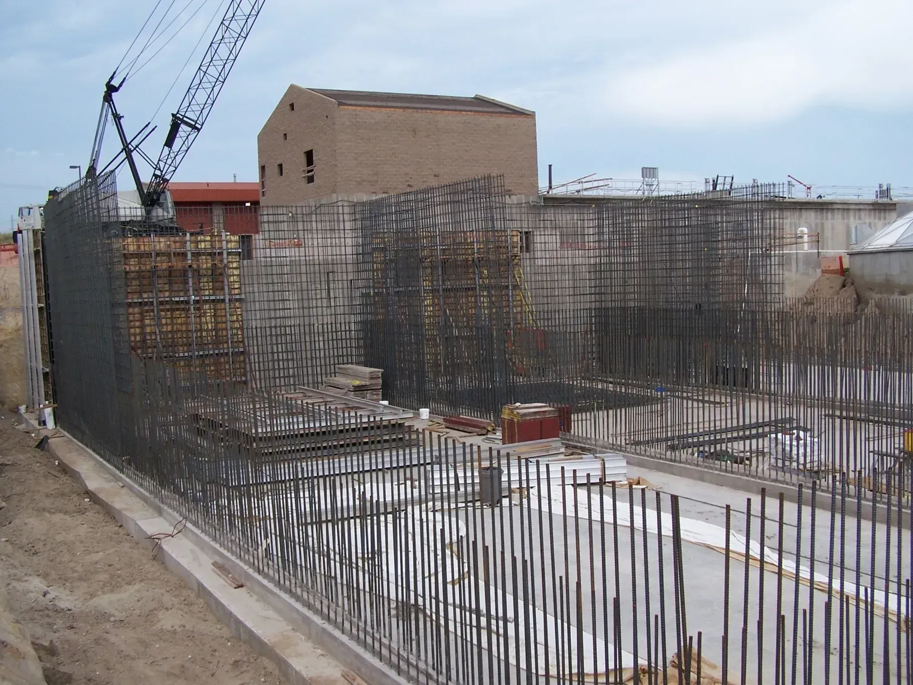 A construction site features exposed rebar grids rising from a concrete foundation, with a brick building in the background.