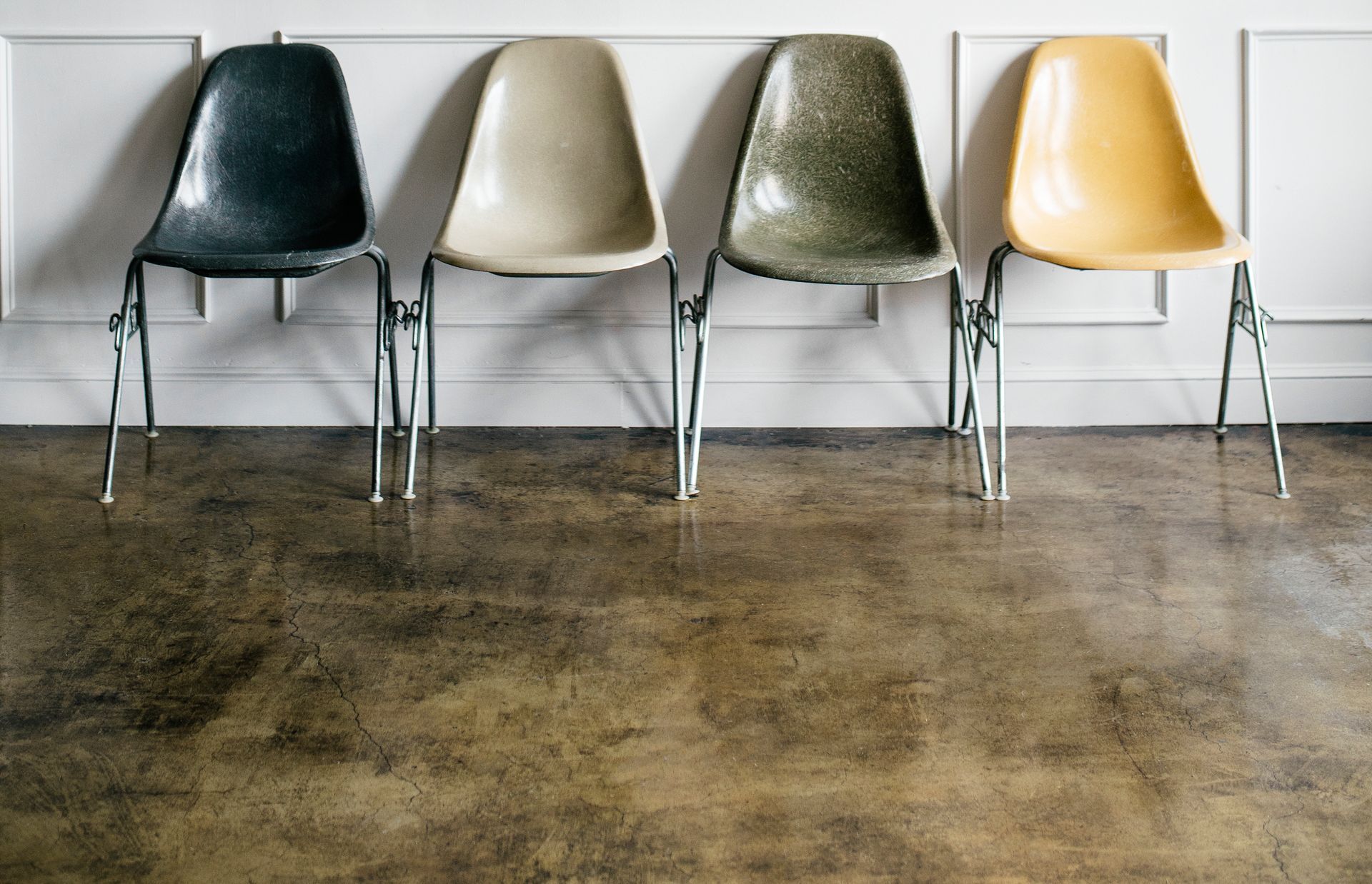 Four mid-century modern style chairs in black, off-white, olive green, and yellow lined up against a white paneled wall.