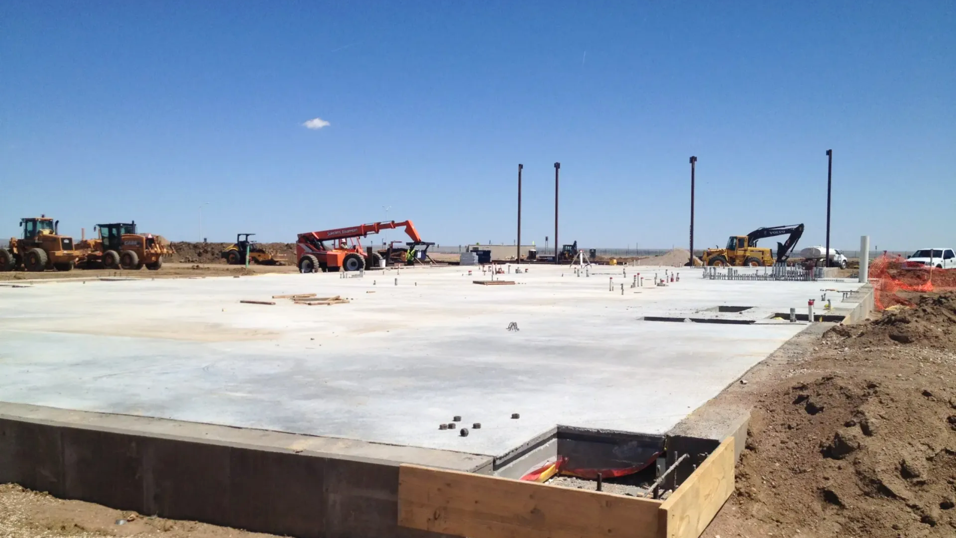 A concrete construction foundation slab under a clear blue sky, surrounded by earth-moving machinery.