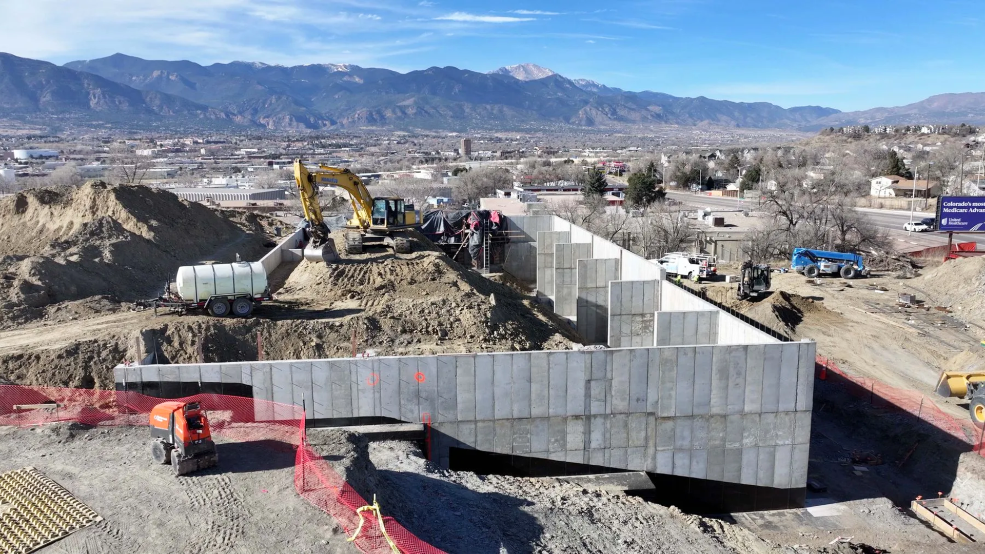 An aerial view of an active construction site with concrete retaining walls, excavators, and mountains in the background.